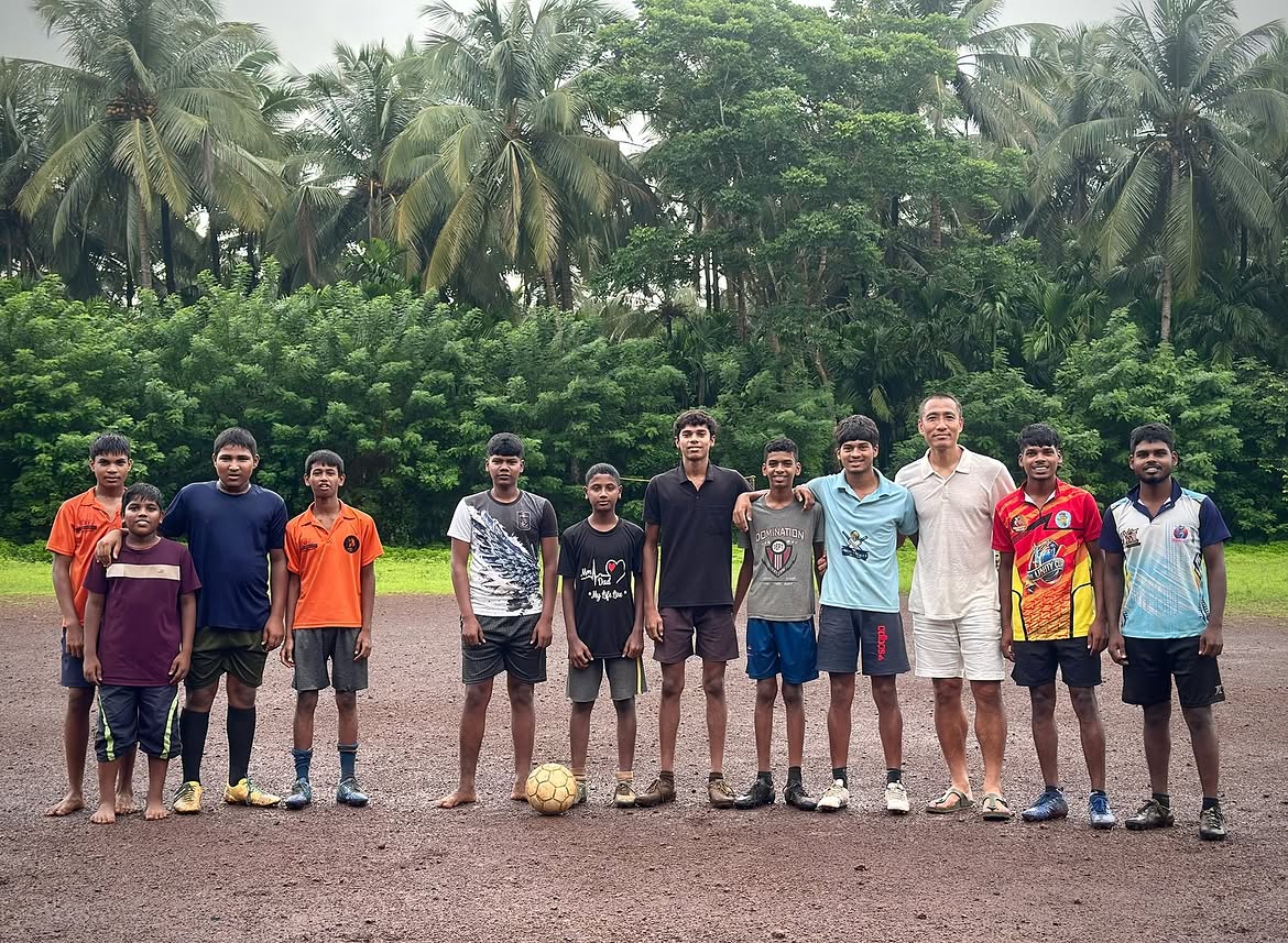 Young footballers in Goa posing for a photo on a rain-soaked pitch surrounded by palm trees